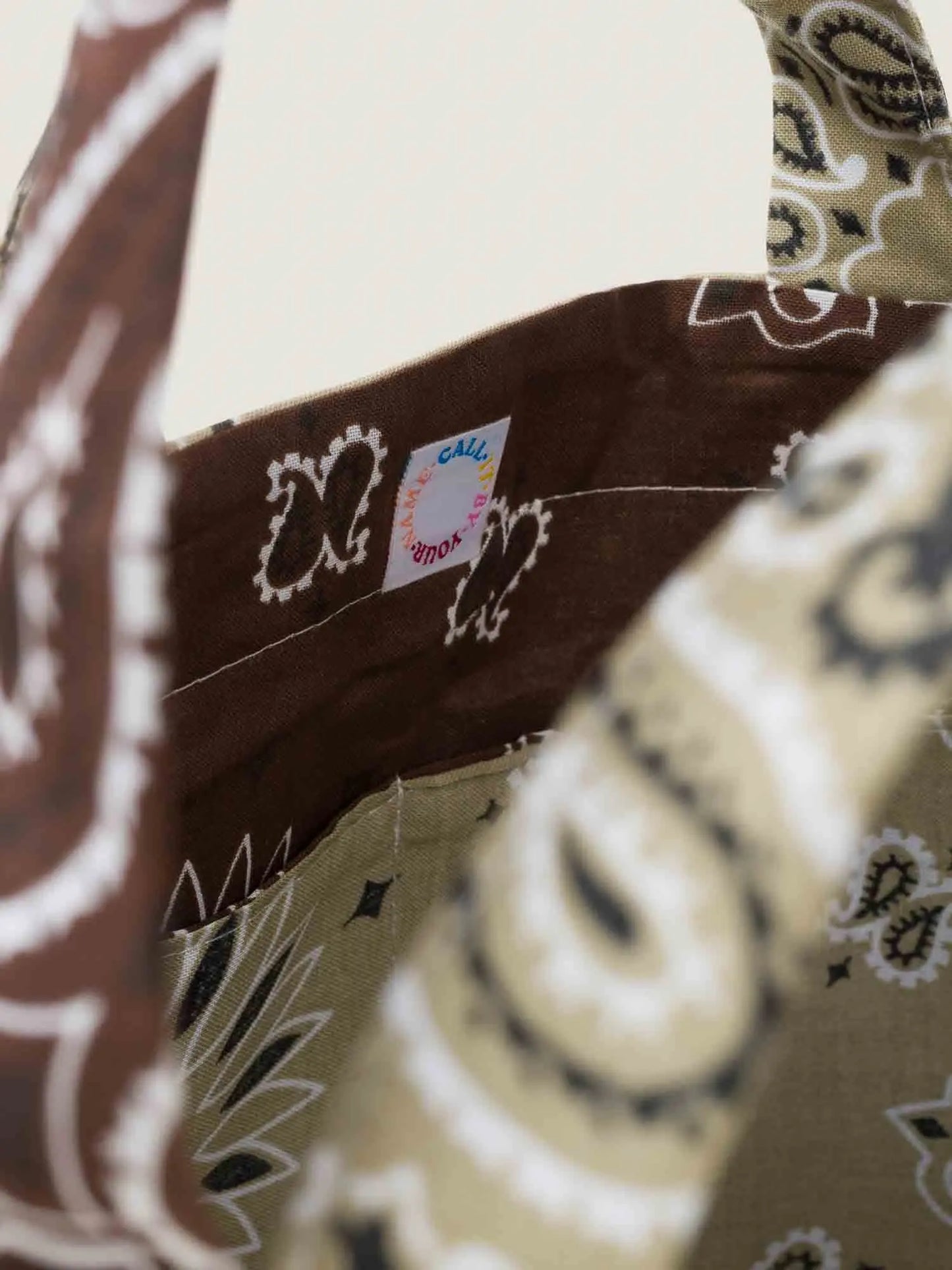 Tote bag with brown bandana pattern on a beige background, with inside and outside pockets, 2 handles and 2 shoulder straps, from brand Call it by your name, close-up