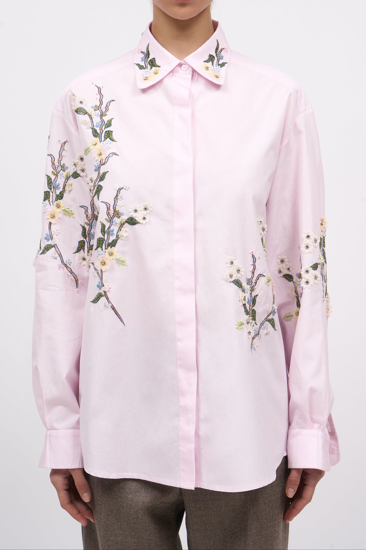 A model wearing a light pink long-sleeve button-up shirt with delicate floral embroidery on the collar, shoulders, and sleeves, paired with brown trousers, shot against a plain light gray background.