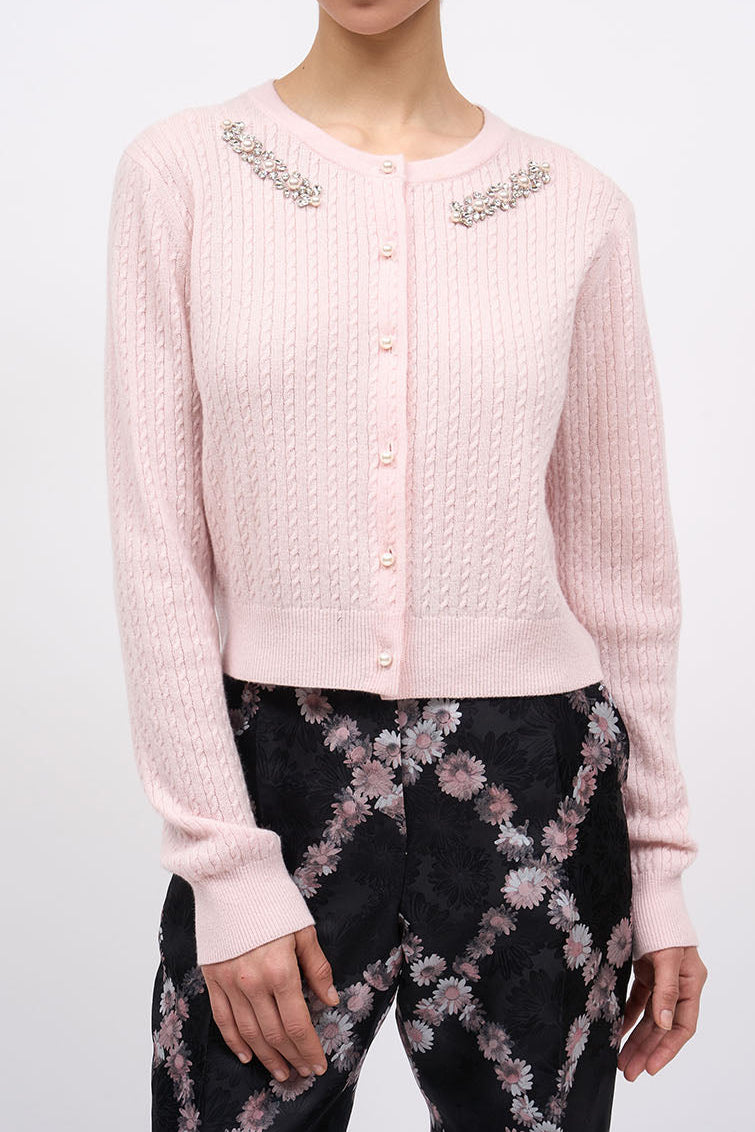 A model wearing a light pink cable-knit cropped cardigan with silver beaded embellishments on the shoulders, paired with black floral print trousers, against a plain light gray background.