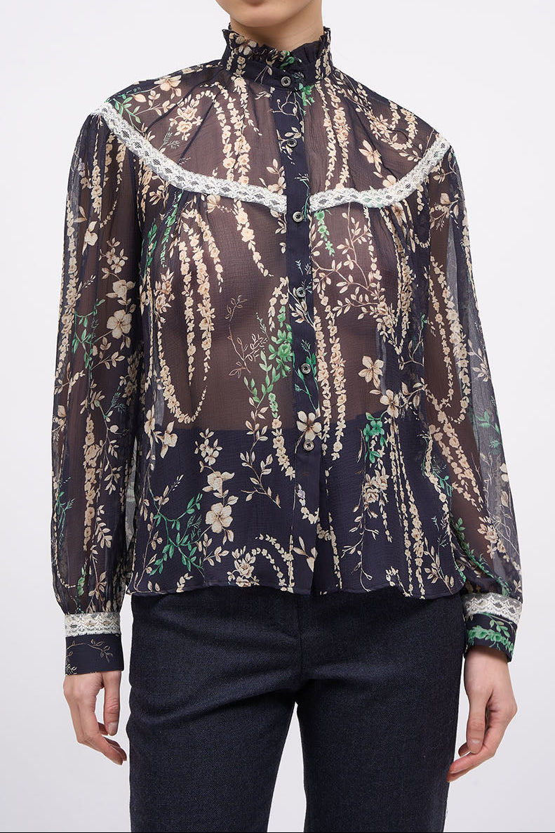 A model wearing a sheer long-sleeve blouse with a dark brown and black paisley pattern, beige floral details, and white lace trim, paired with dark pants, against a light gray background.