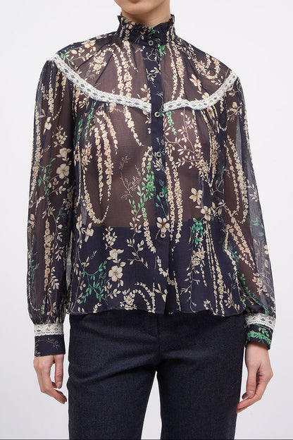 A model wearing a sheer long-sleeve blouse with a dark brown and black paisley pattern, beige floral details, and white lace trim, paired with dark pants, against a light gray background.