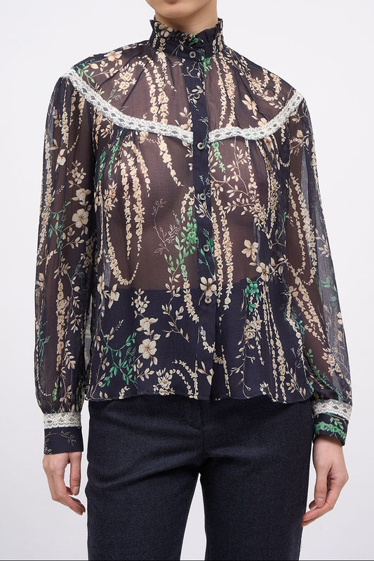 A model wearing a sheer long-sleeve blouse with a dark brown and black paisley pattern, beige floral details, and white lace trim, paired with dark pants, against a light gray background.